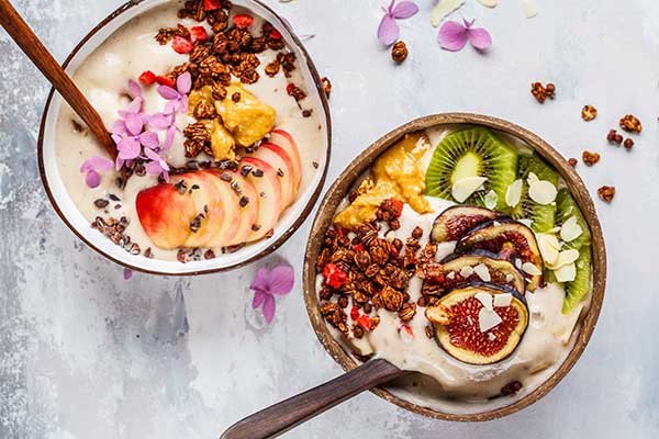 Two coconut bowls containing creamy smooties topped with fresh kiwi figs apples peanut butter and small pink flowers. Image appears on a marble background
