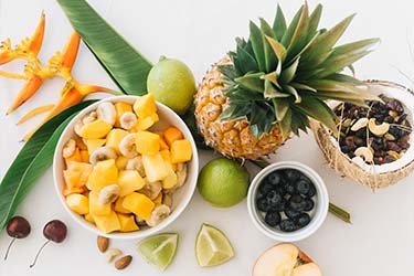 flat lay image of fruits on a white table frozen bananas and mangos in a white bowl and some dried fruit and nuts in a coconut