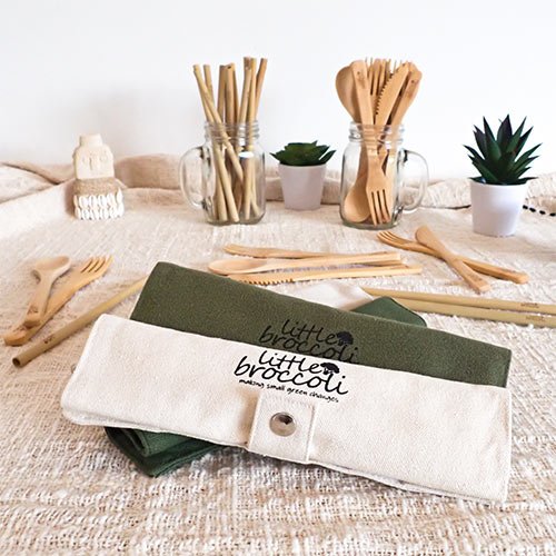 A stack of little broccoli bamboo cutlery pouches on a cream fabric with loose bamboo cutlery and straws laying around it. There are some glasses of cutlery and plants in the background.