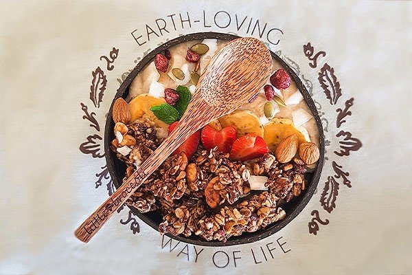 A close up top view image of a colourful breakfasy bowl containing fresh fruits and granola. A reclaimed palm wood spoon is placed on top of the bowl. The background reads 'earth loving way of life'